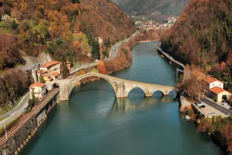 Il **Ponte del Diavolo**, con le sue arcate in pietra, attraversa un fiume circondato da colline autunnali e case.