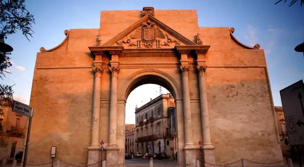 Porta Napoli a Lecce, un maestoso arco in pietra illuminato dal tramonto, accoglie i visitatori con la sua architettura barocca.