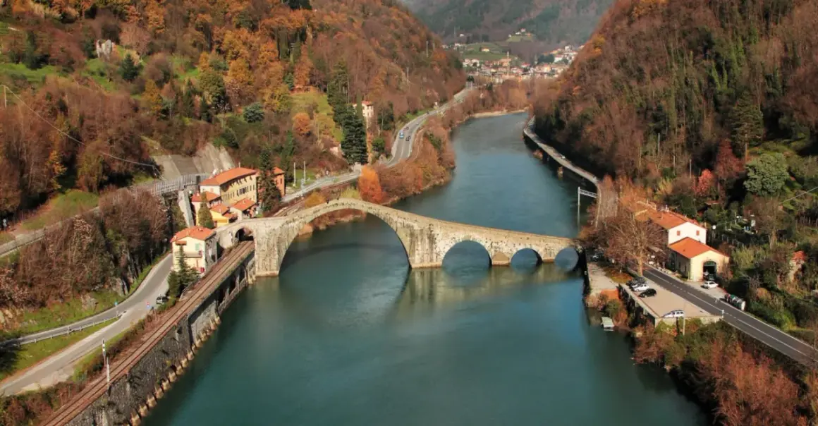 Il **Ponte del Diavolo**, con le sue arcate in pietra, attraversa un fiume circondato da colline autunnali e case.