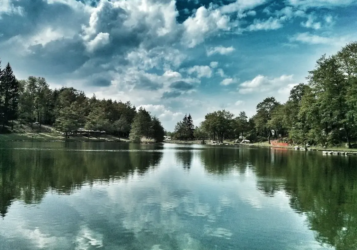 Un lago tranquillo circondato da alberi verdi, con nuvole drammatiche nel cielo. Un'oasi di pace tra i laghi dell'Emilia Romagna.