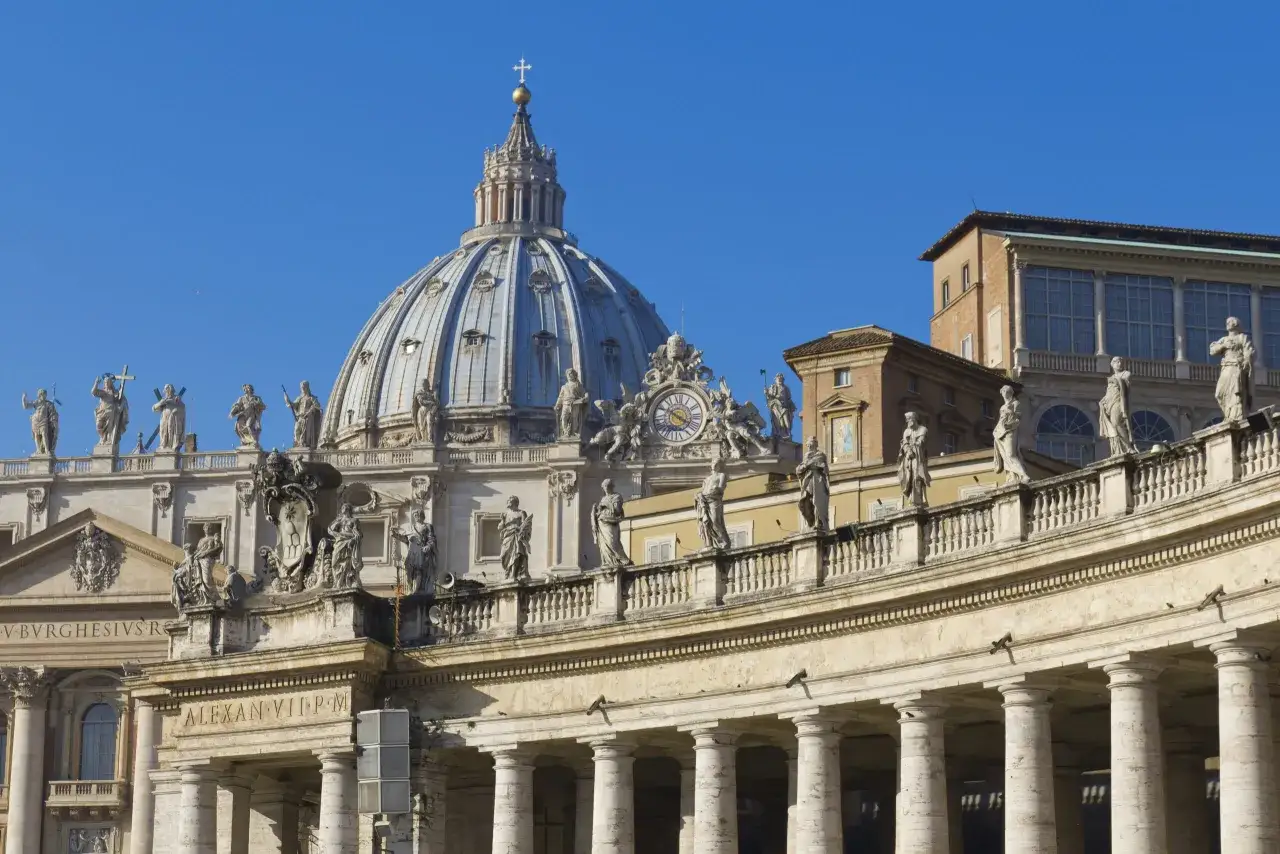 La maestosa cupola della chiesa di San Pietro a Roma domina il cielo azzurro, circondata da statue e colonnati.