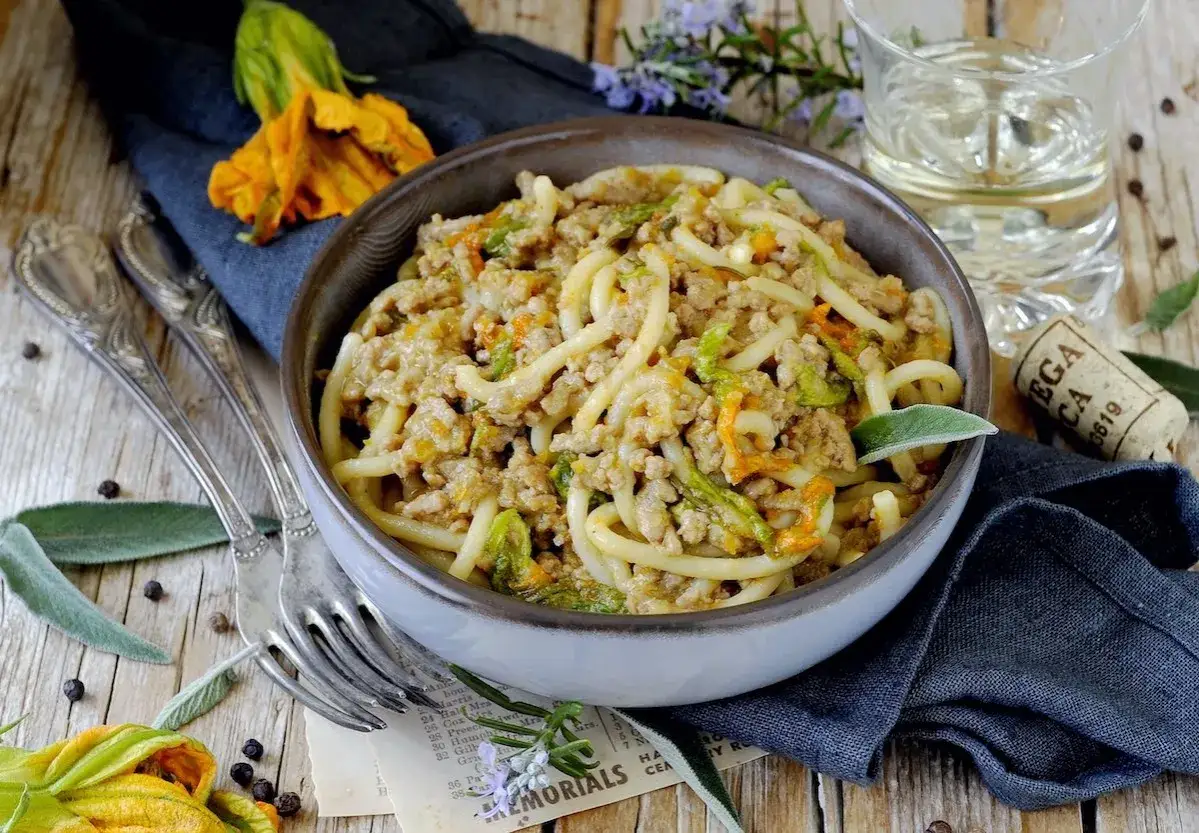 Pici al ragù con fiori di zucca, un piatto rustico e saporito, servito in una ciotola su un tavolo di legno.