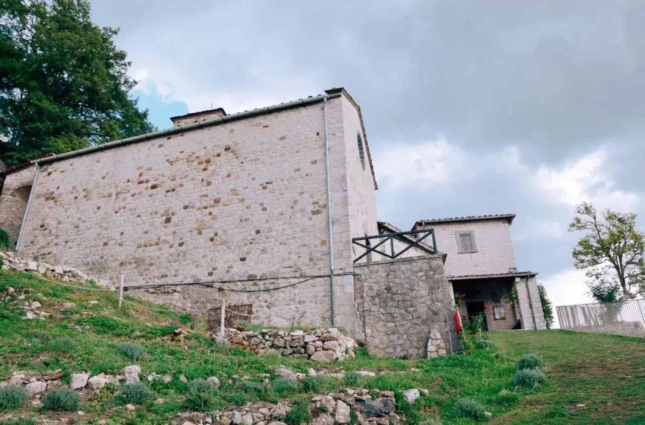 Edifici in pietra con tetto spiovente e un piccolo balcone in legno. Il eremo di Cerbaiolo si erge su un pendio erboso, con alberi sullo sfondo.
