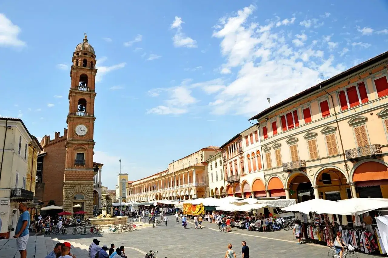 Piazza gremita a Faenza, cosa vedere: torre civica, portici eleganti e mercato vivace sotto un cielo azzurro.