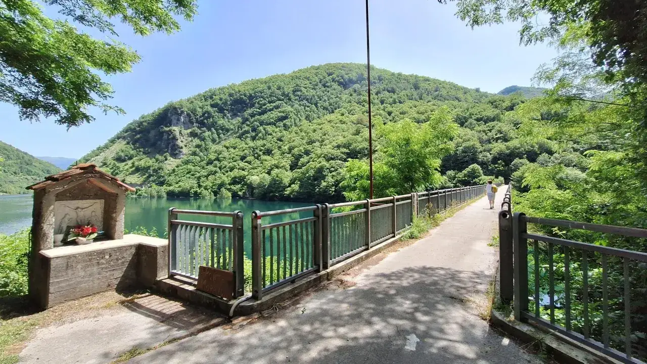 Un piccolo edicola votiva con fiori, un ponte con ringhiera e una persona che cammina, con montagne verdi e un lago sullo sfondo. I vagli di sotto offrono una vista mozzafiato.