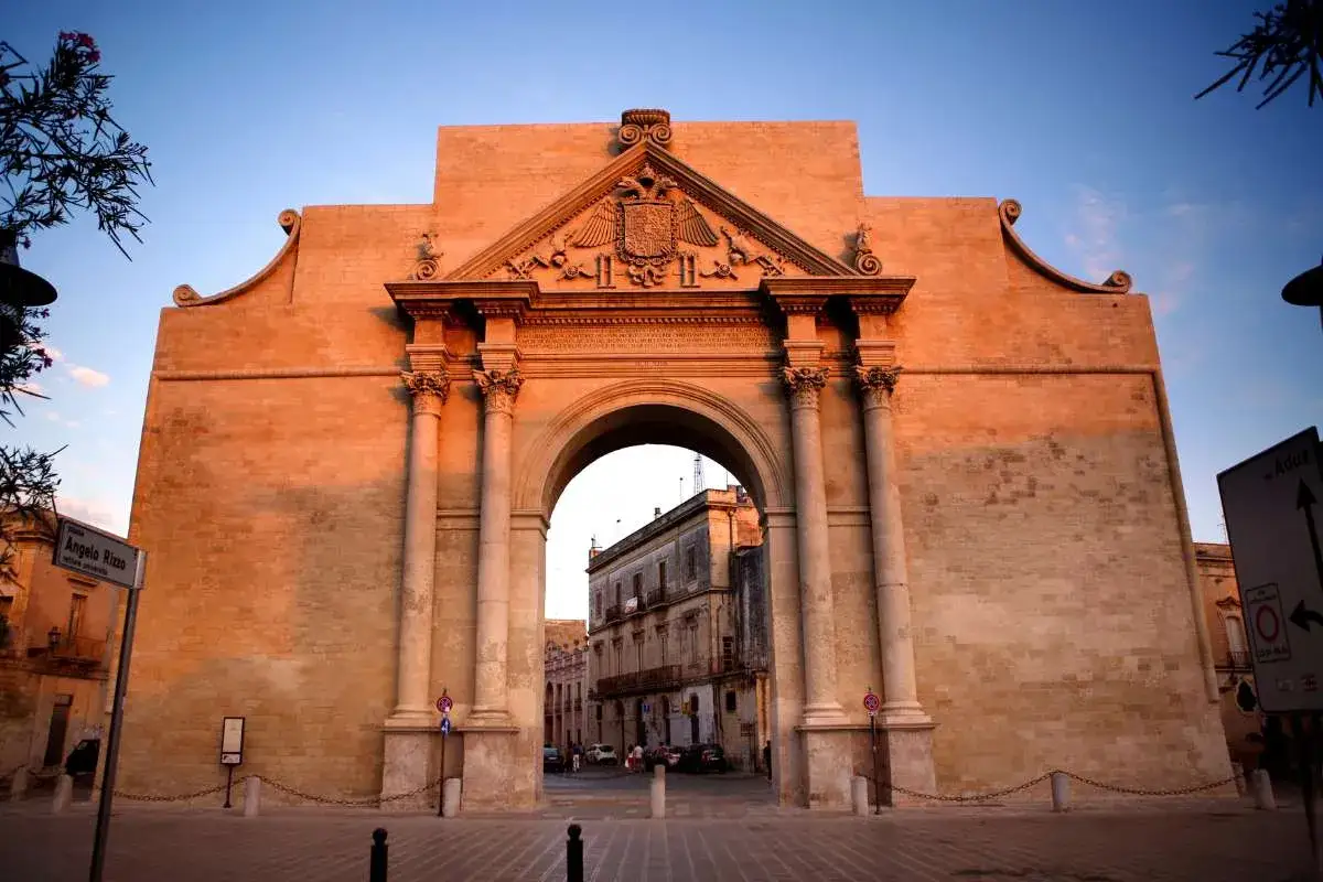 Porta Napoli a Lecce, un maestoso arco trionfale in pietra illuminato dal sole del tramonto, accoglie i visitatori.