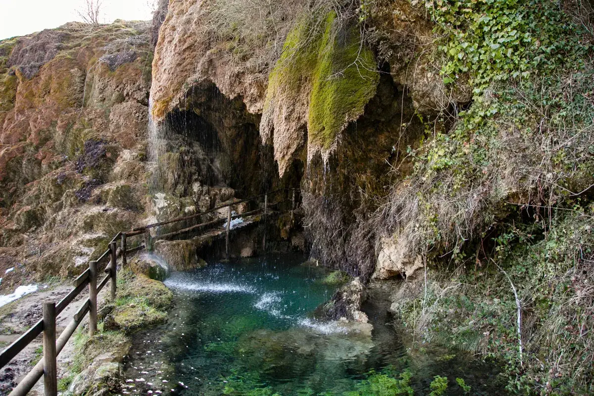 Cascata che scende tra rocce muschiose, formando una pozza d'acqua cristallina. Un sentiero con staccionata in legno conduce alle grotte di Labante, un luogo incantevole.