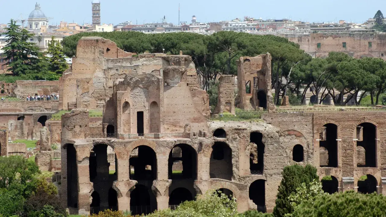 Imponenti rovine del Foro Romano Palatino, con archi e mura in mattoni che si stagliano contro il cielo azzurro e il verde degli alberi.