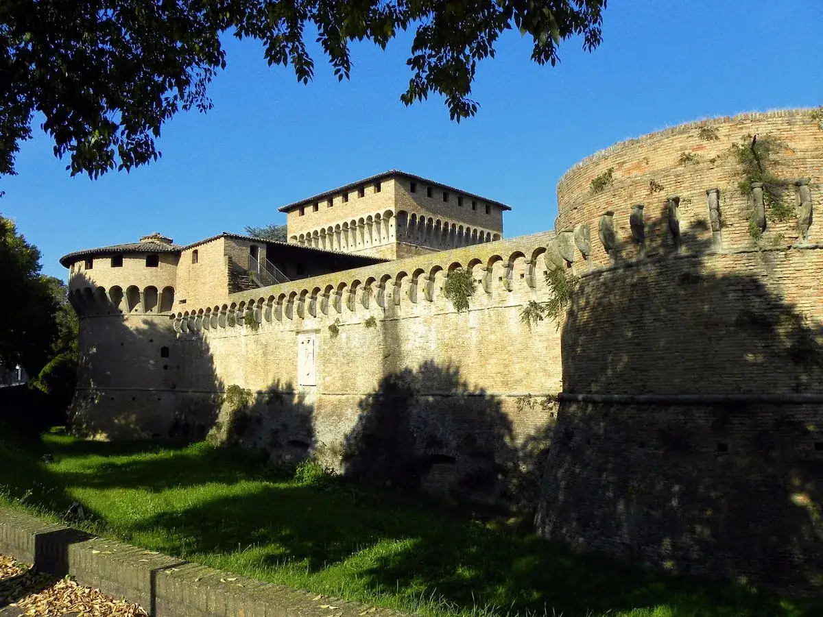 Rocche e mura imponenti, un esempio di cosa vedere a Forlì, con torri merlate e un fossato verde sotto il cielo azzurro.