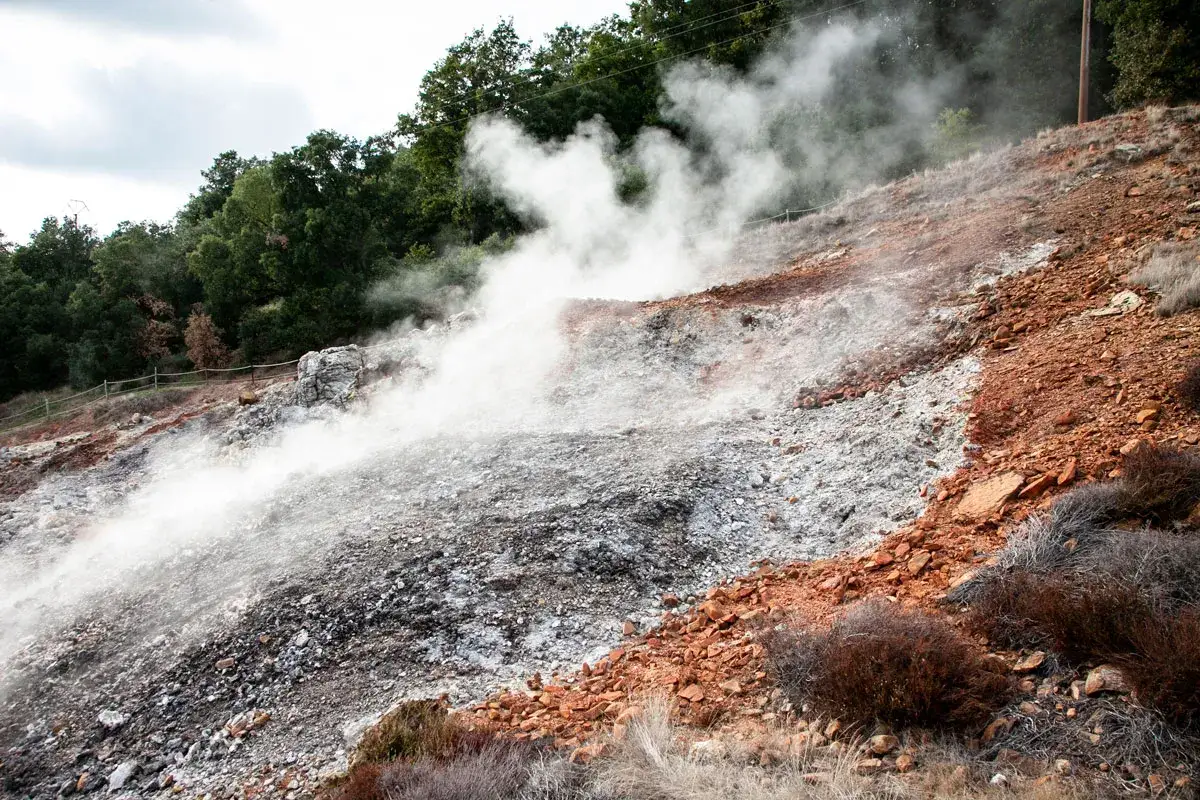 Fumarole attive nel parco delle Biancane, con vapore che sale dal terreno roccioso e argilloso, circondato da vegetazione e alberi.