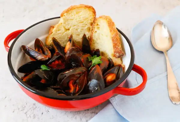 Un invitante guazzetto di cozze in una pentola rossa, accompagnato da pane croccante e una forchetta.
