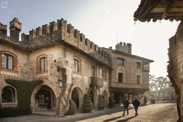 Antico borgo Visconti a Piacenza, con torri merlate e archi in pietra. Due uomini passeggiano nel cortile illuminato dal sole.